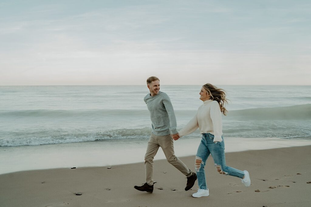 An engaged couple runs on the beach along the water during their engagement session at the Indiana Dunes.  They both wear cozy fall outfits.
