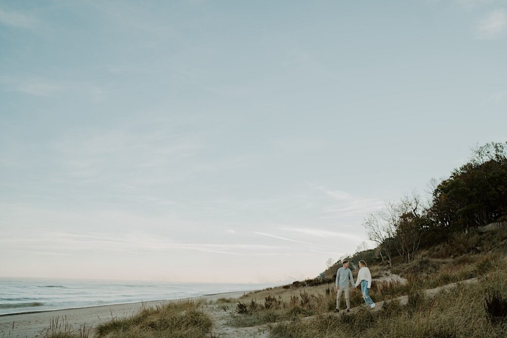 An engaged couple walks through the dunes at the Indiana Dunes during their fall engagement session at sunset.