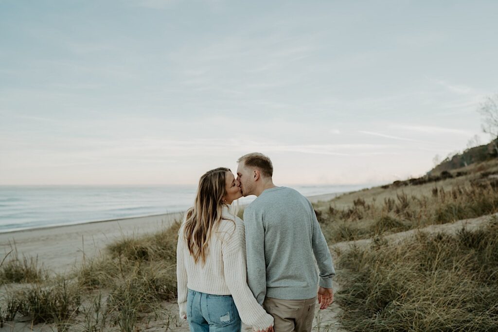 An engaged couple kisses and walks away from the camera while holding hands in the dunes in Indiana