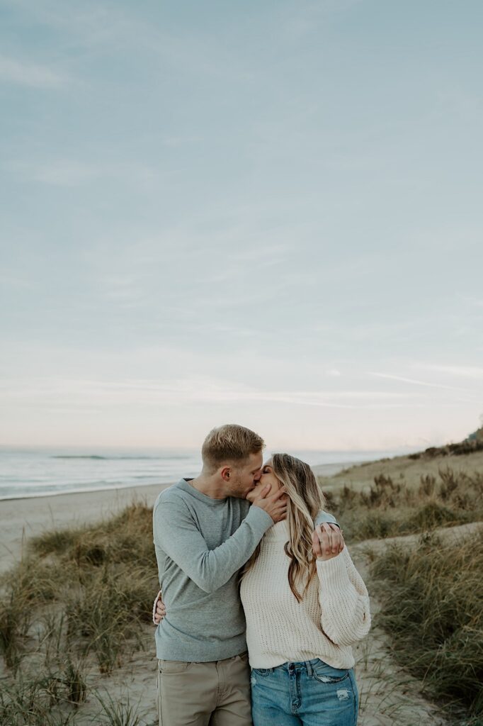 An engaged couple kisses and walks towards the camera while holding hands in the dunes in Indiana