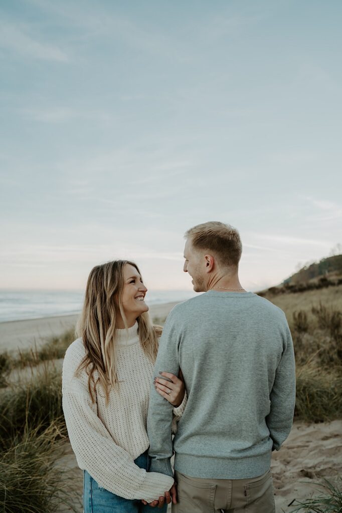 A fiancée holds her soon to be husbands arm and faces the camera while smiling at him while he faces his body away from the camera smiling at her.