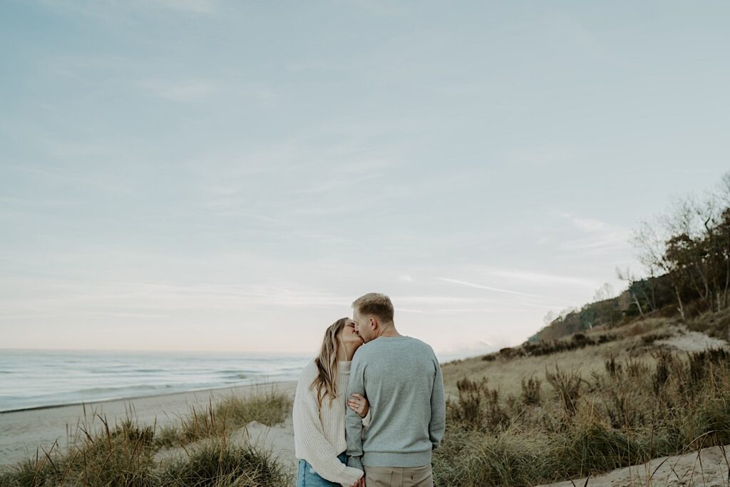 An engaged couple kisses while standing on the beach at the Indiana Dunes State Park.  They are wearing fall sweaters and the sky is light blue with a little bit of pink.