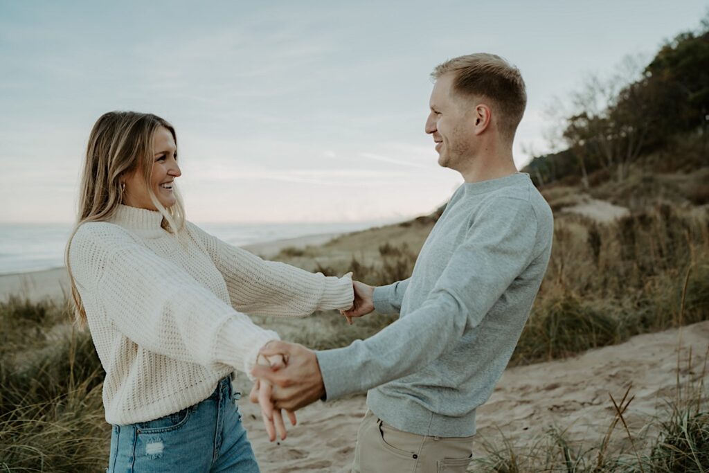 A couple holds both hands and faces each other while smiling at one another.  They are standing on the beach at the Indiana Dunes State Park.