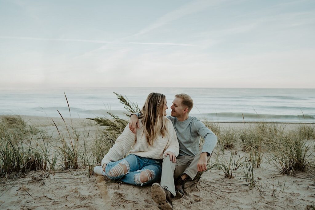 A couple sits in the tall grasses on the beach, snuggling close to one another during their engagement session at the Indiana Dunes State Park.