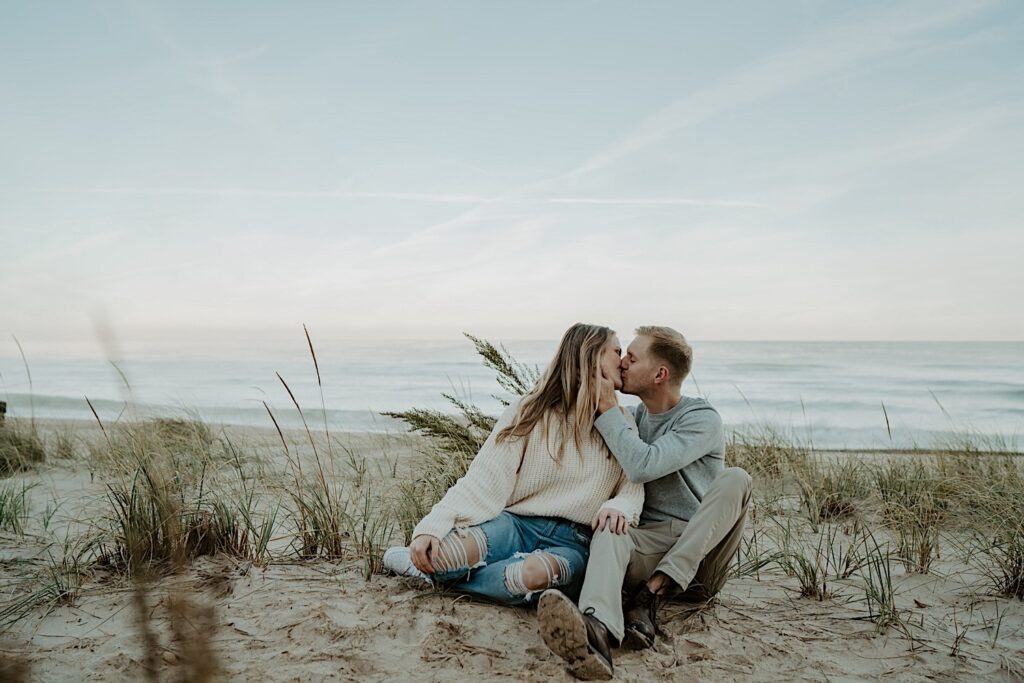 A couple sits in the tall grasses on the beach, snuggling close to one another and kissing during their engagement session at the Indiana Dunes State Park.