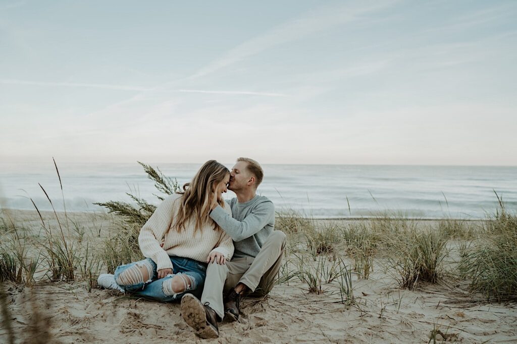 A couple sits in the tall grasses on the beach, snuggling close to one another and kissing one another during their engagement session at the Indiana Dunes State Park.