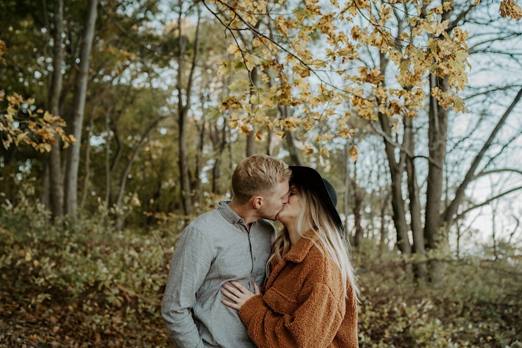 A couple stands under a tree with yellow leaves in cozy fall outfits during their engagement session in Indiana Dunes State Park