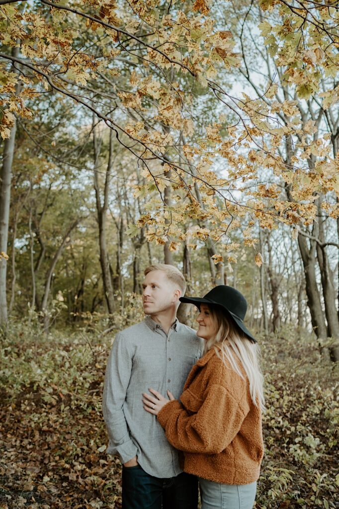 A couple stands under a tree with yellow leaves in cozy fall outfits during their engagement session in Indiana Dunes State Park
