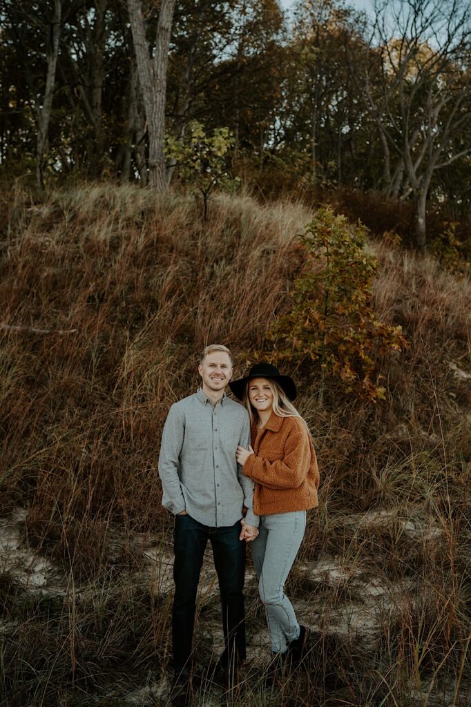A couple stands in front of brown and green colored grasses in the Indiana Dunes during their engagement session.