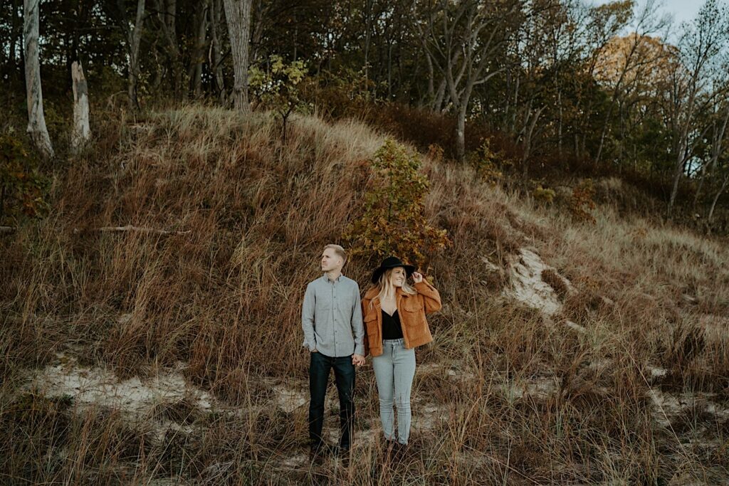 A couple stands while holding hands and looking away from one another surrounded by tall grasses.  The fiancée wears an orange colored sherpa jacket, light jeans and a black flat brimmed hat.  The fiancé wears dark colored jeans and a light grey button up.