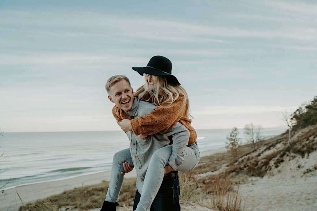 A fiancée rides on her fiancé's back laughing during their engagement session.  You can see Lake Michigan in the background and a bit of the Indiana Dunes.