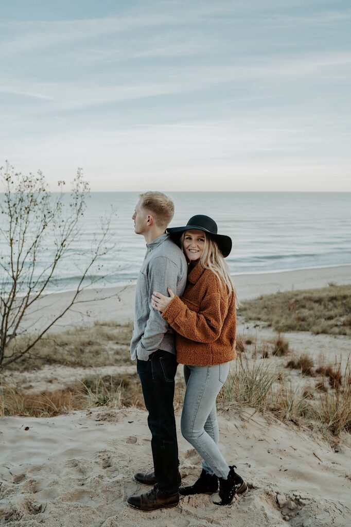 A fiancée stands up against her fiancé's back while holding his bicep and smiling at the camera.  He looks off into the distance, they are both standing on the beach.