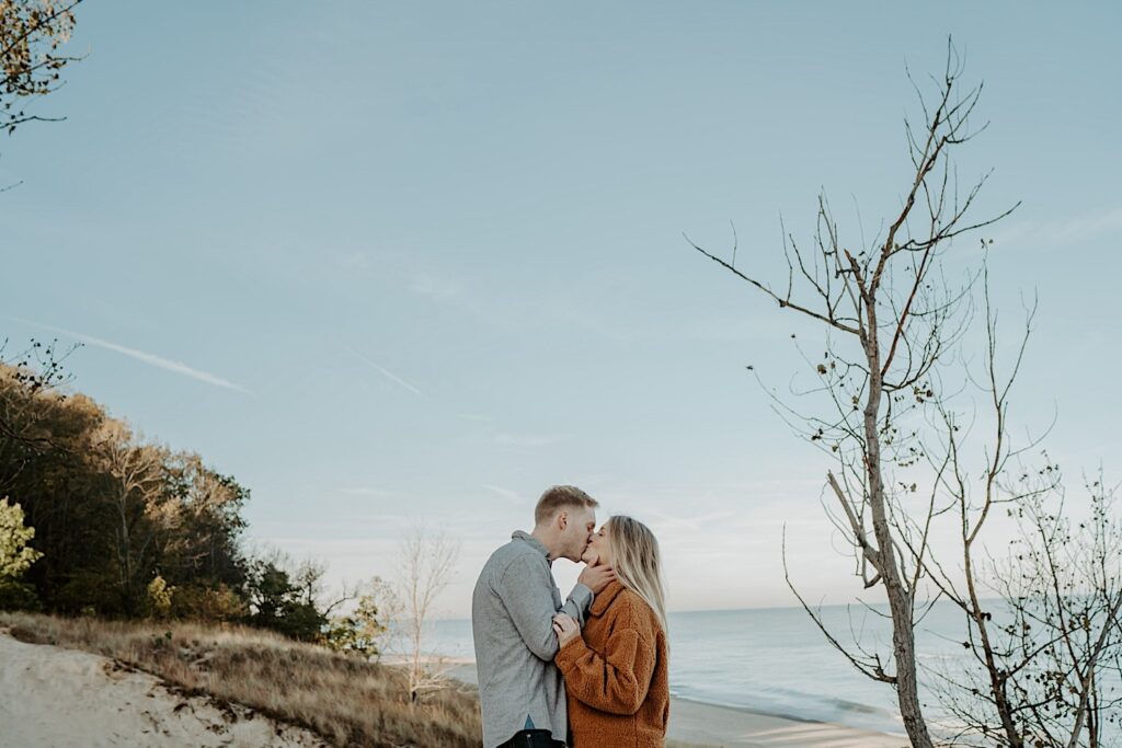 An engaged couple kisses during their engagement session at the Indiana Dunes.  You can see the cloudless blue sky in the background along with the Indiana Dunes and Lake Michigan.