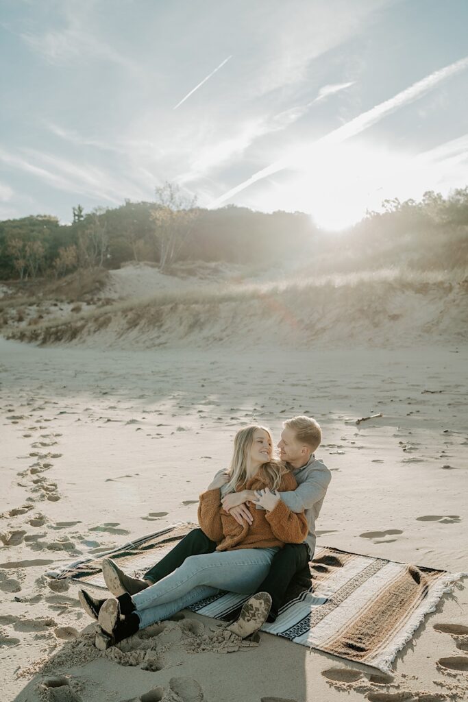 An engaged couple sits on a striped blanket snuggling at the Indiana Dunes during their engagement session.