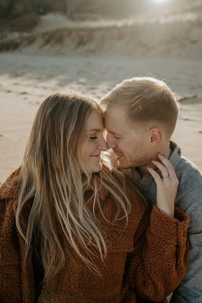 An engaged couple sits with their noses pressed together while they smile on the beach during their Indiana engagement session.
