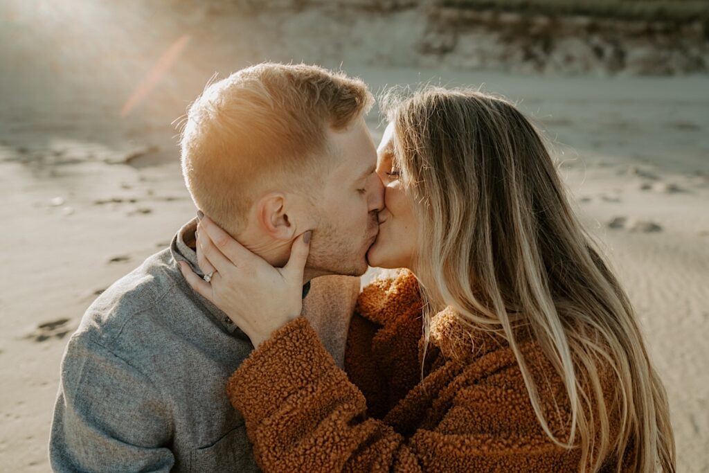 An engaged couple sits together on the beach kissing during their Indiana engagement session at sunset.
