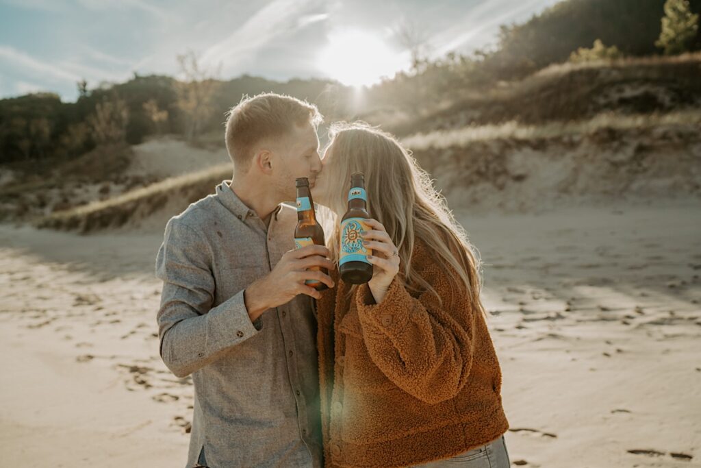 An engaged couple clinks their Oberon beer bottles while walking towards the camera and kissing during their session.