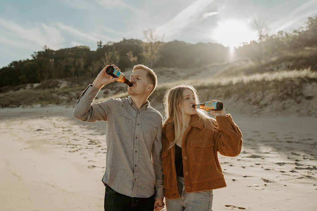 An engaged couple holds hands on the beach in the fall while sipping out of their Oberon bottles.