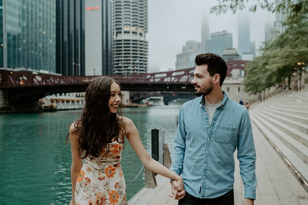 An engaged couple walks along the Chicago River smiling at one another during their engagement session.