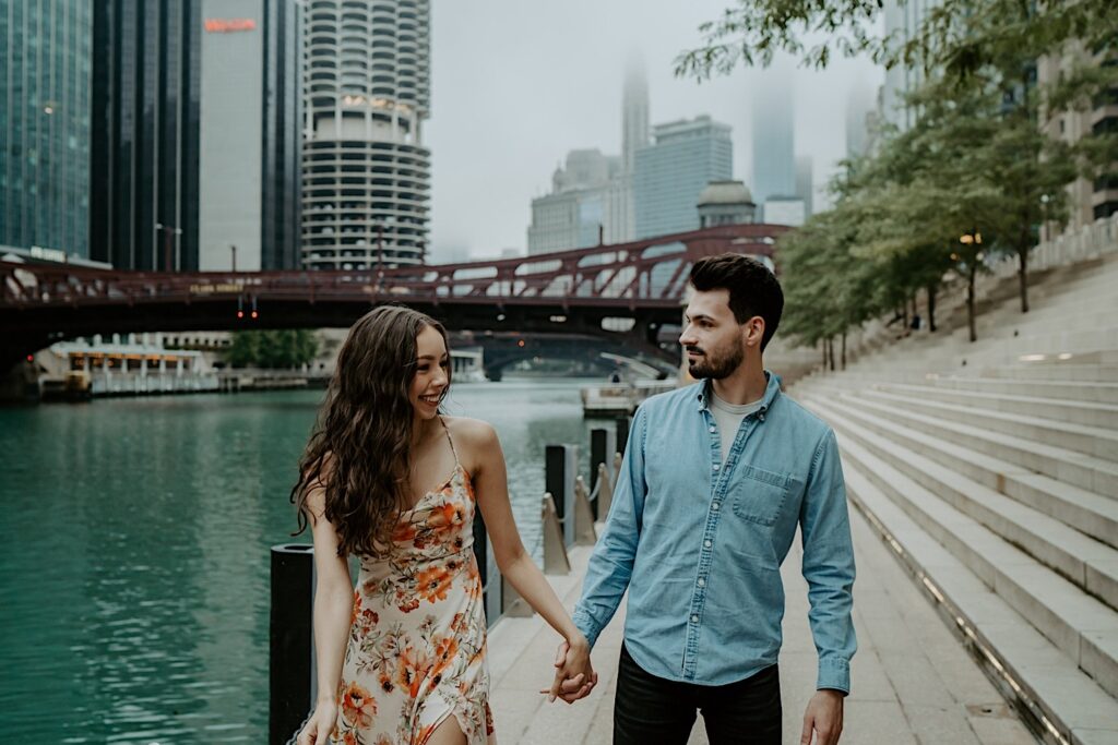 An engaged couple walks along the Chicago Riverwalk smiling at each other and holding hands.