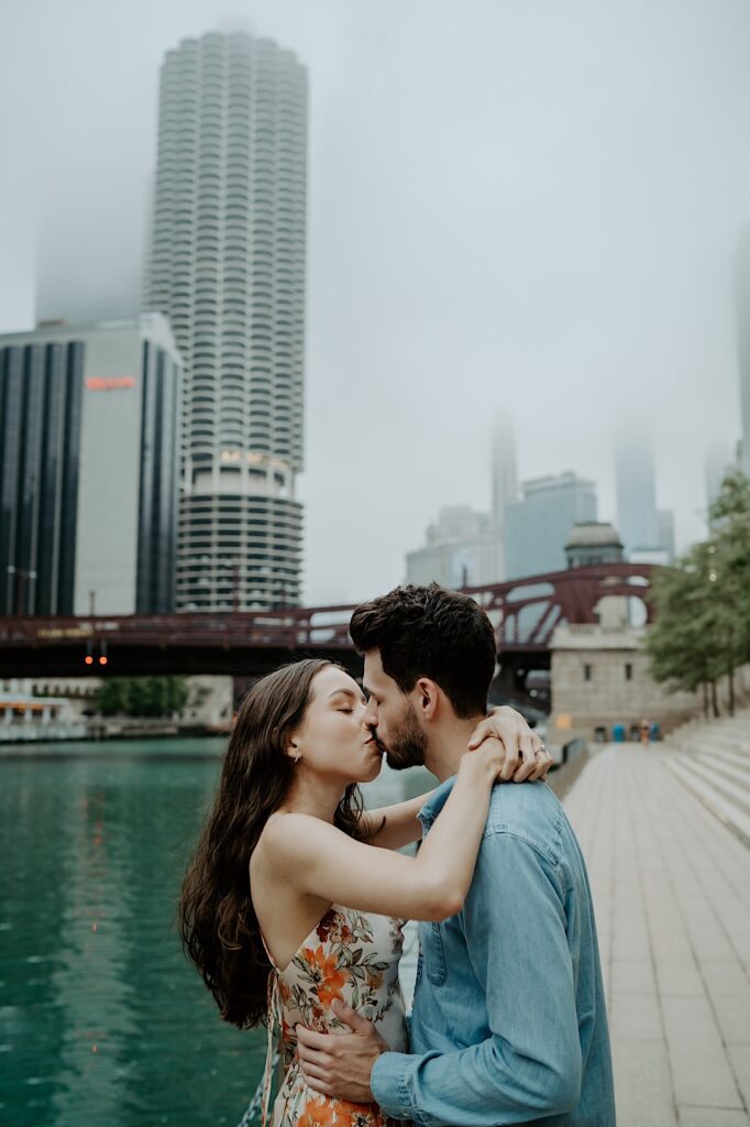 A fiancée holds his fiancé around her waist and she puts her arms around his shoulders while kissing during their engagement session on the Chicago River.