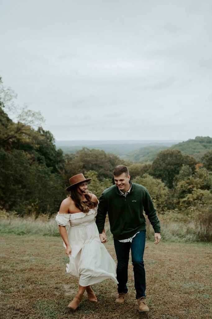 An engaged couple walks while holding hands and looking at one another heading towards the camera with a beautiful green backdrop