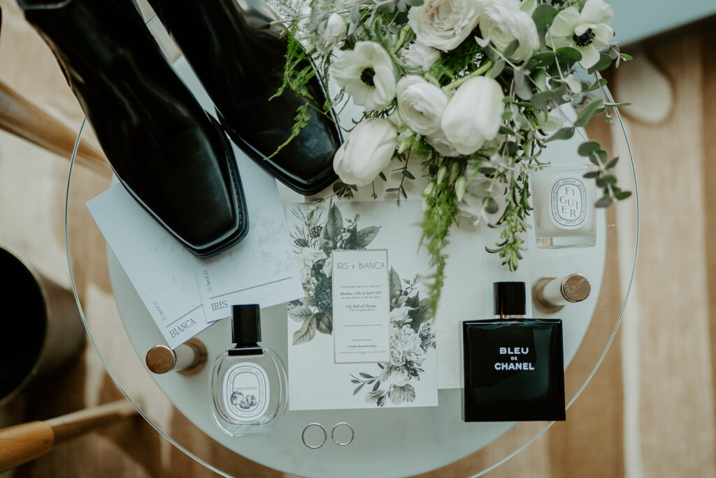 A full wedding day flat lay including the couples custom vow books with their names on them, one of the brides chelsea square toe black wedding boots, two different perfumes, silver wedding bands, and white flowers, mostly roses, and tulips.