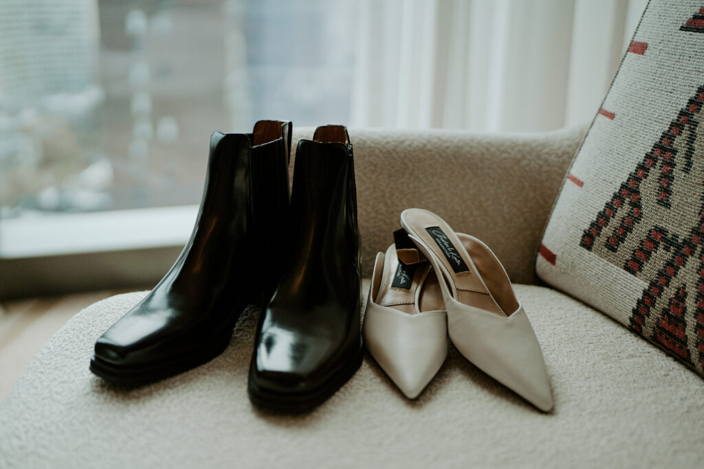 Both sets of the brides shoes sitting next to a window letting in a lot of natural lighting.  One set of bridal shoes is a pair of black square toed chelsea boots and the other pair is a pair of low nude heals.