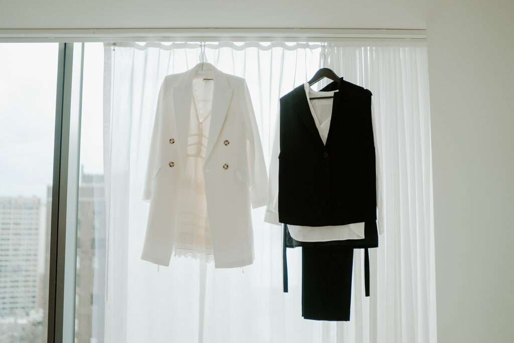 The bridal attire for both brides on their wedding day hanging in front of a white translucent curtain.  One bride is wearing a white lace dress with a white blazer and the other bride is wearing black pants, a white button up and a white vest.