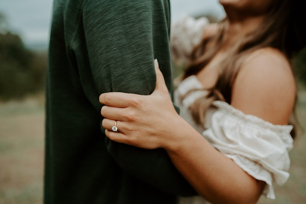 A close up image of the soon to be brides engagement ring, as she holds her fiancé's bicep tight.  He's wearing a green shirt that matches the greens throughout the state park.