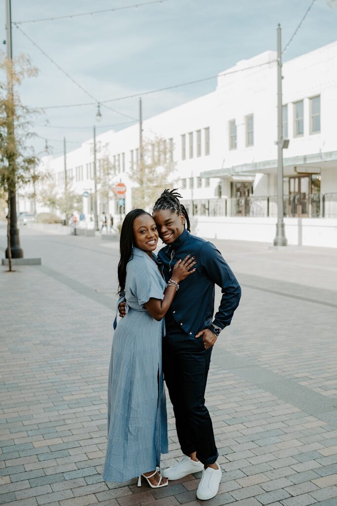 An engaged couple hugs and smiles at the camera while standing on a brick sidewalk in downtown Indianapolis
