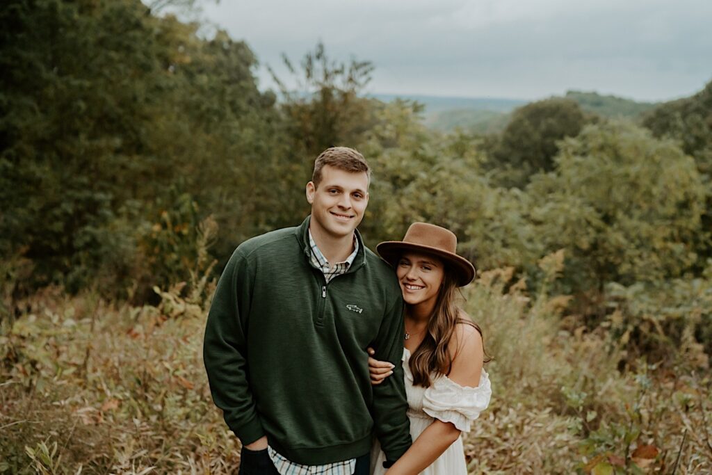 Fiancé's hug each other tight while smiling at the camera during their engagement session.  One wears a cream dress and a brown brimmed hat while the fiancé wears a green quarter zip and khaki's