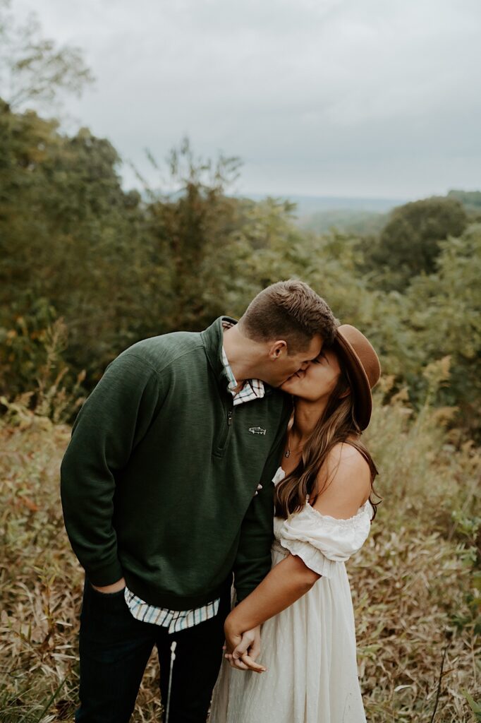 Fiancé's hug each other tight while kissing during their engagement session.  One wears a cream dress and a brown brimmed hat while the fiancé wears a green quarter zip and khaki's.