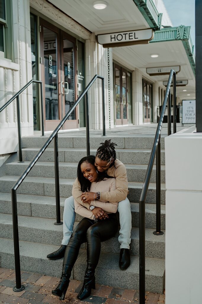 A couple hugs each other while sitting on marble steps together outside the Bottleworks hotel during their engagement session in Indianapolis.