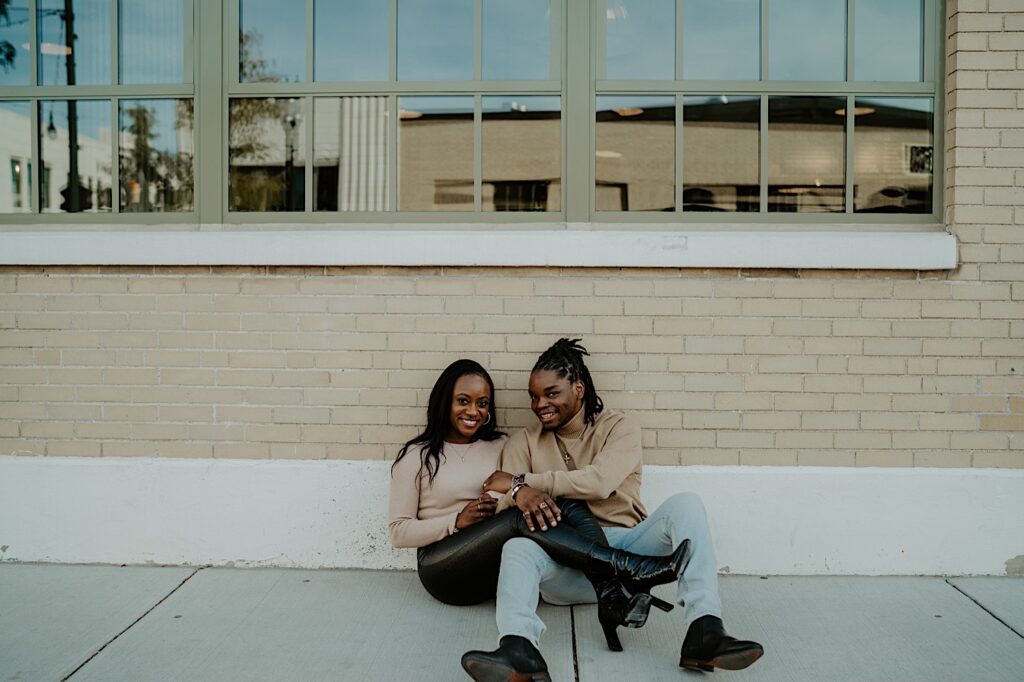 An engaged couple sits on the ground in front of a beige brick wall while smiling at the camera