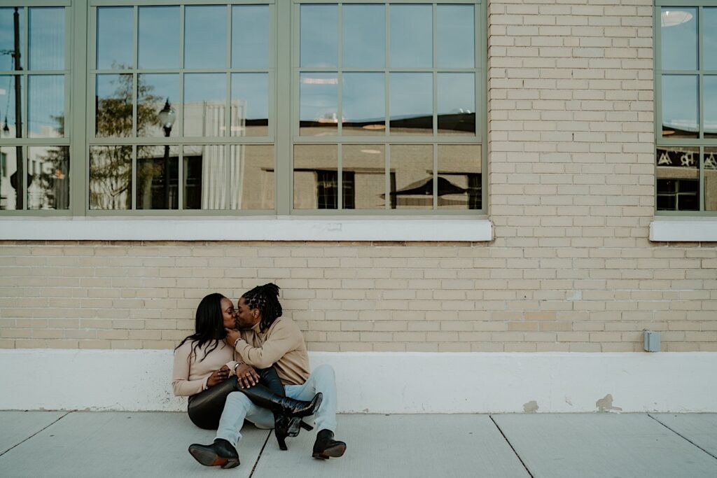 An engaged couple sits on the ground in front of a beige brick wall in downtown Indy while kissing.