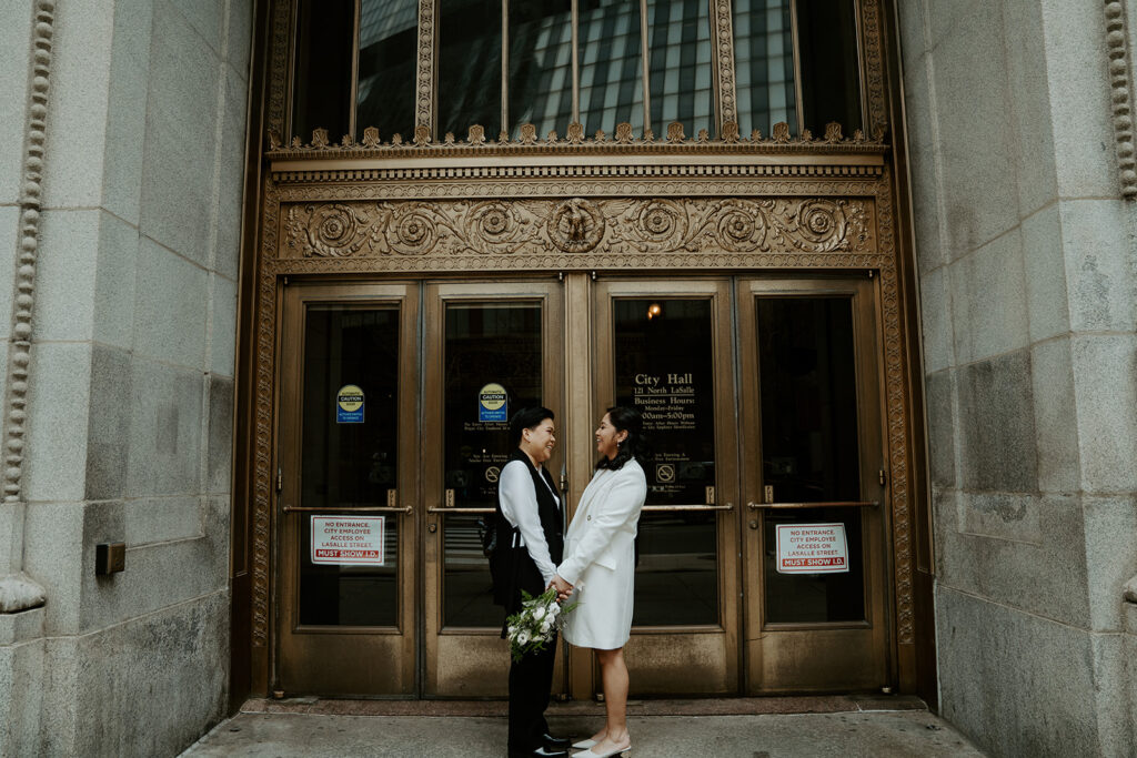 Brides stand outside of Chicago City Hall in front of the iconic ornate gold doors, holding hands and smiling at one another.