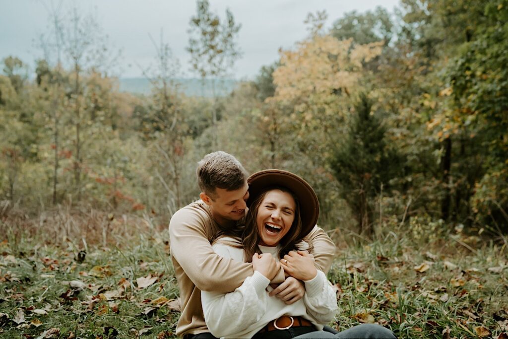 Fiancé's both wear beige tops with denim and the soon to be bride wears a wide brimmed hat while smiling and sitting in the grass at Brown County State Park