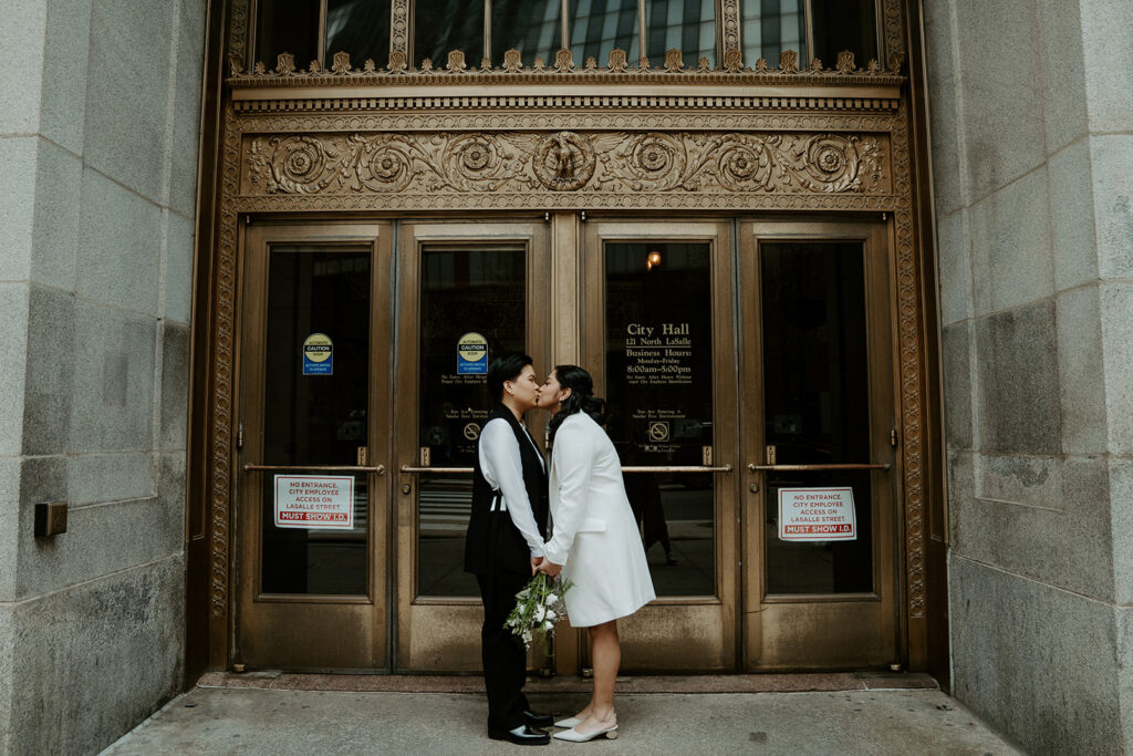 Brides stand outside of Chicago City Hall in front of the iconic ornate gold doors, holding hands and kissing one another.