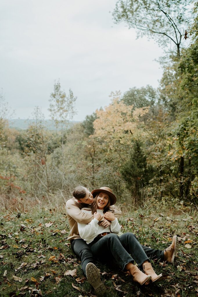 An engaged couple sits on the ground while holding each other and smiling.