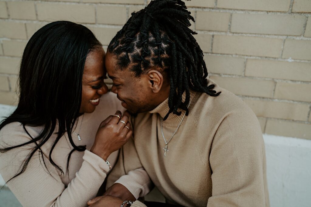 A couple presses their foreheads together almost kissing while sitting in front of a beige wall while wearing beige outfits.
