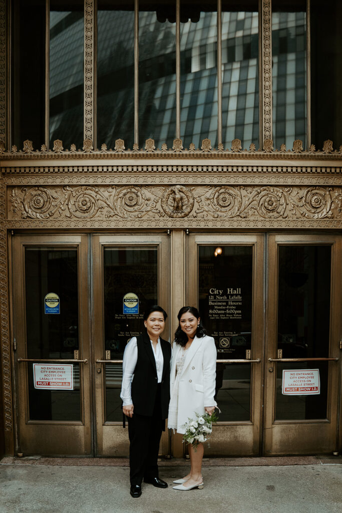 Brides stand outside of Chicago City Hall in front of the iconic ornate gold doors, holding hands and smiling at the camera.