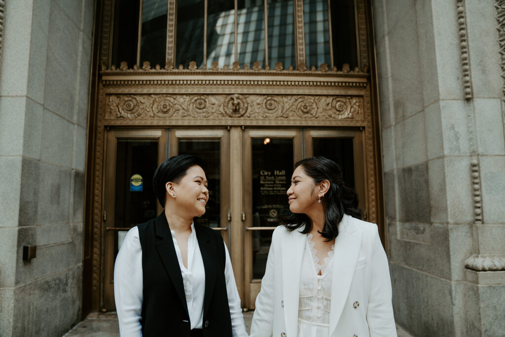Brides stand outside of Chicago City Hall in front of the iconic ornate gold doors, holding hands and smiling at one another.