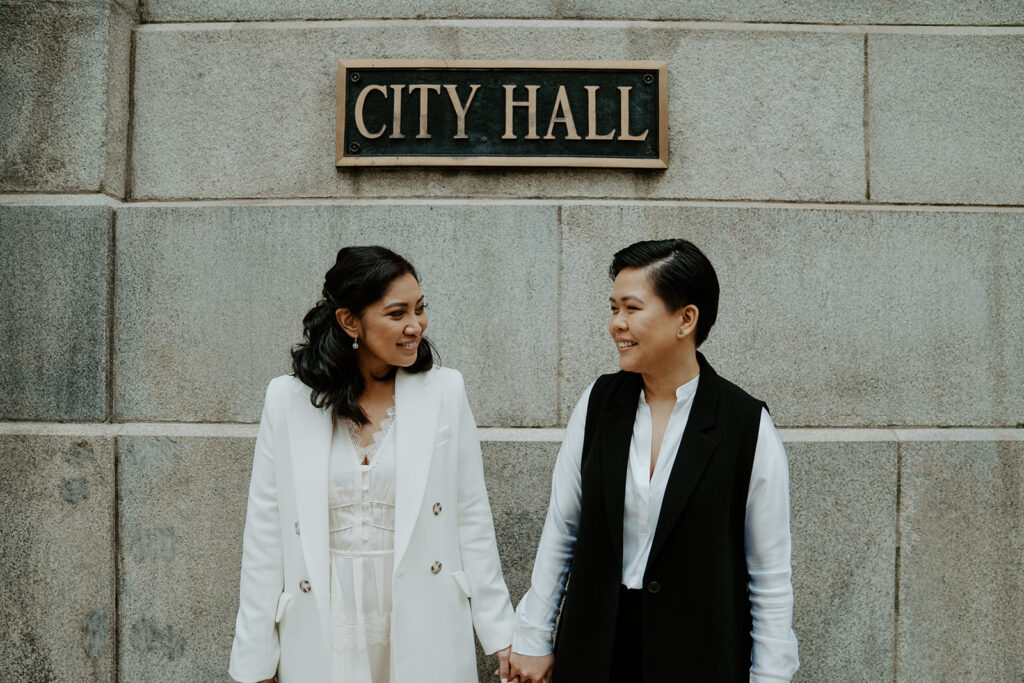 Brides stand in front of the City Hall sign at the Chicago's City Hall immediately following their wedding ceremony.  They are holding hands and looking at one another.