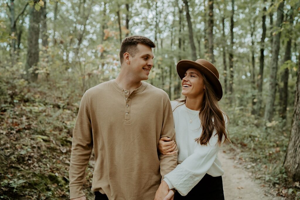 An engaged couple holds hands while smiling at each other and walking towards the camera during an engagement session in Indiana in the forest