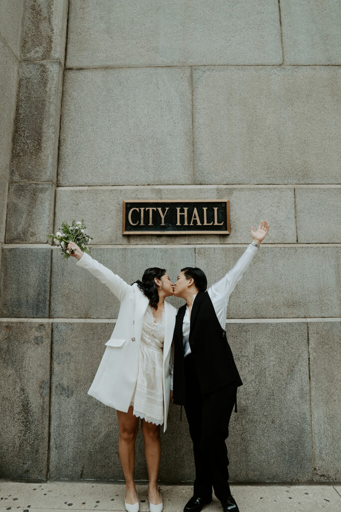 Brides stand in front of the City Hall sign at the Chicago's City Hall immediately following their wedding ceremony.  They are kissing and holding their hands up in celebration.
