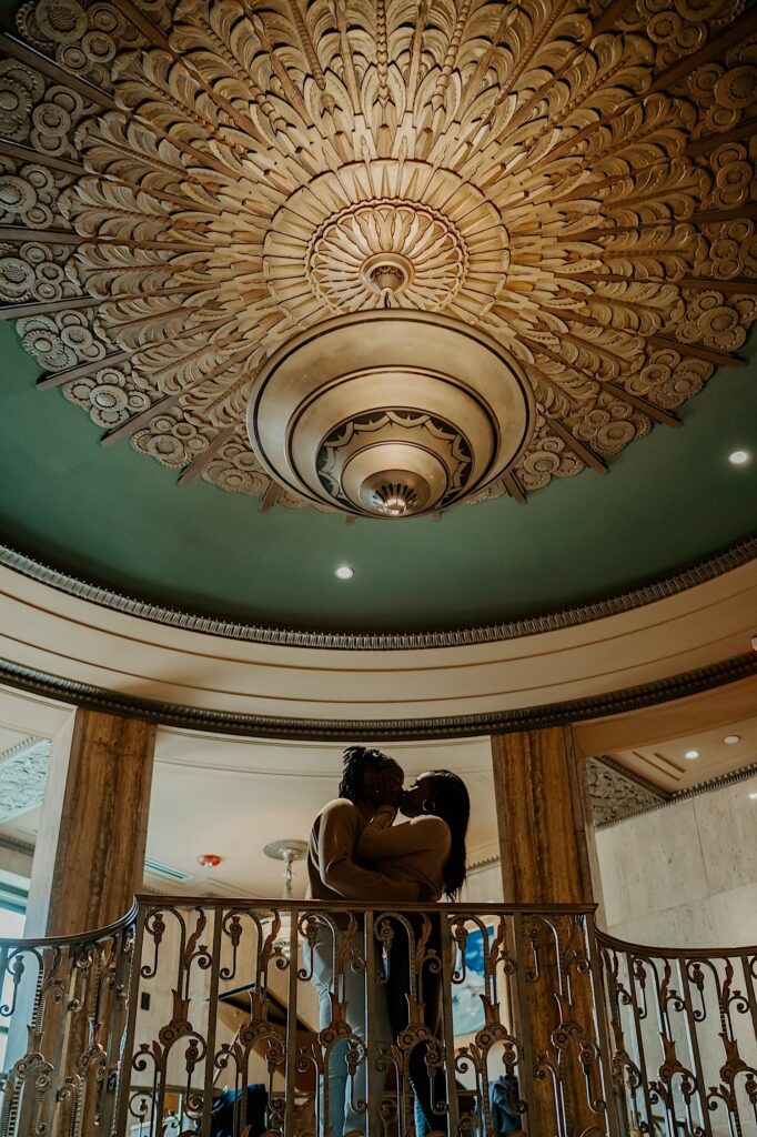 A couple kisses while looking over a gold balcony at the Bottleworks Hotel during their engagement session.