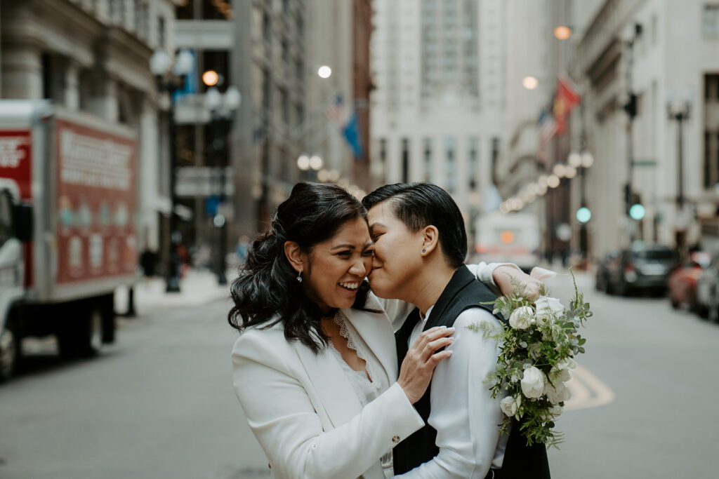 Newly weds stand in the middle of a street in Chicago sharing an intimate moment with one another.  One bride kisses her bride on the cheek