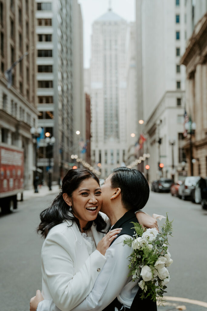 Newly weds stand in the middle of a street in Chicago sharing an intimate moment with one another.  One bride kisses her bride on the cheek
