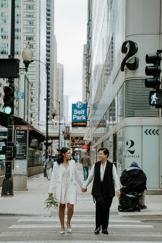 Two brides hold hands after their elopement walking across the street in the Loop in Chicago.